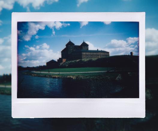 A medieval castle. In front a green field of grass. Behind a blue sky and clouds floating by.