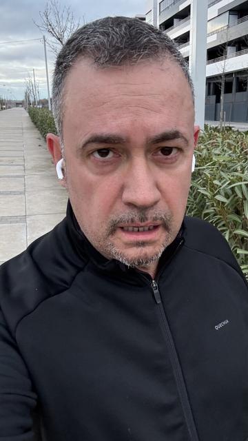 Selfi de un hombre con piel clara y cabello corto y canoso, caminando al aire libre en una zona urbana. Lleva auriculares inalámbricos blancos y viste una chaqueta deportiva negra con cremallera de la marca Quechua. Su expresión muestra esfuerzo, con la boca ligeramente abierta y la mirada fija en la cámara. Detrás de él hay edificios modernos, arbustos y un paseo peatonal con árboles sin hojas, bajo un cielo nublado.