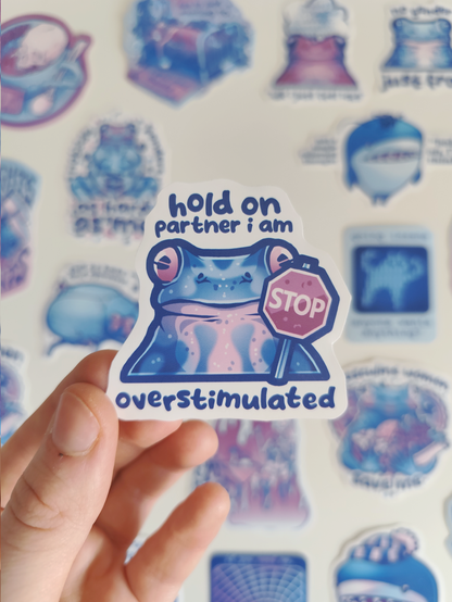 hand holding up a sticker of a frog with a stop sign titled "hold on partner i am overstimulated"
