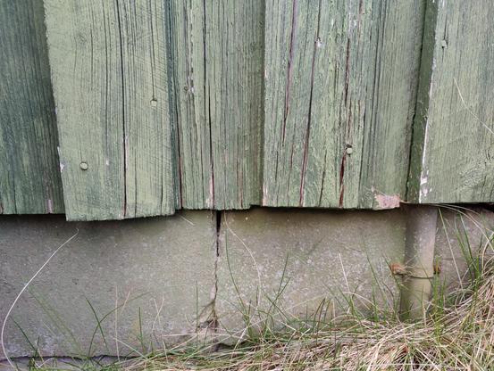 The lower edge of a green wooden facade. It is old, cracked and uneven, but not rotten.