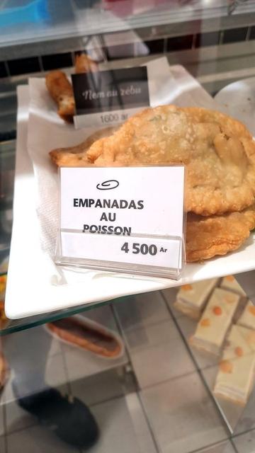 A close-up photo of a display case in a grocery store in Antananarivo, Madagascar, showing a white plate with golden-brown fried fish empanadas. A sign in front of the plate reads "EMPANADAS AU POISSON" with a price of "4 500 Ar.