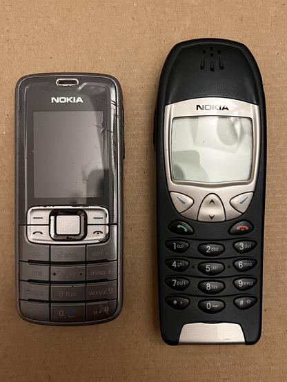 Zwei Nokia Mobil Telefone der frühen 2000er Jahre, aus der Vor-Smartphone Zeit. Rechts das ältere und etwas größere Nokia 6210, links das kleinere Nokia 3109c, welches einem Nokia 3110 Classic ohne Kamera entspricht. Beide Handys sind ausgeschaltet und liegen auf einen hellbraunen Pappuntergrund.