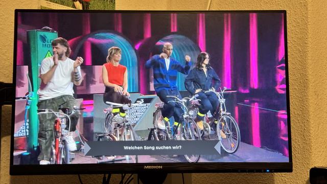 A TV screen displays four contestants sitting on bicycles, engaged in a game show. They are smiling and participating while colorful stage lights illuminate the background. A prompt asking "Welchen Song suchen wir?" (Which song are we looking for?) appears at the bottom
