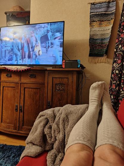 A TV showing MASH, on top an oak cupboard, and a pair of legs wearing grey knee socks, resting on a sofa