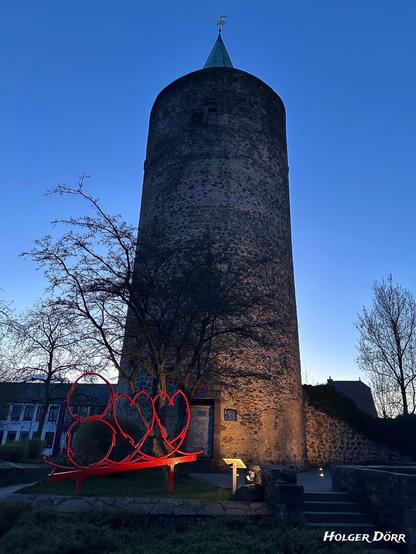 Der Diebesturm in Grünberg bei einsetzender Dämmerung. Der runde, mittelalterliche Turm aus Bruchstein mit spitzem Dach erhebt sich vor einem tiefblauen Abendhimmel.
An seiner Basis steht eine rote, moderne Kunstinstallation in Form von drei stilisierten Herzen, die auf einer geneigten Plattform ruhen. Der Turm und die Umgebung werden von warmem Licht angestrahlt, wodurch ein stimmungsvolles Zusammenspiel zwischen alter Architektur und moderner Kunst entsteht. Im Vordergrund führen Stufen und ein Weg zur Turmöffnung, daneben eine Infotafel mit Beleuchtung. Kahle Bäume umrahmen die Szene.