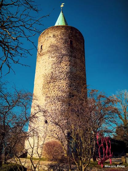 Der Diebesturm, ein mittelalterlicher Rundturm aus grobem Naturstein, ragt in den strahlend blauen Himmel. Sein grünes Spitzdach mit einer goldenen Wetterfahne verleiht ihm einen markanten Abschluss. Umgeben von kahlen Asten und einem modernen roten Kunstwerk im Vordergrund, steht er als stiller Zeuge der Zeit.