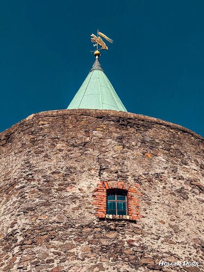 Eine Nahaufnahme des oberen Teils des Diebesturms zeigt das grüne Spitzdach mit der goldenen Wetterfahne in Form eines geflügelten Wesens. Darunter ein kleines, vergittertes Fenster mit rot umrahmten Backsteinen, das sich in die raue Steinmauer einfügt.
Der tiefblaue Himmel bildet einen eindrucksvollen Kontrast zur historischen
Architektur.
