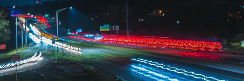 Abstract long exposure of nighttime traffic on Interstate 80 near Coralville, Iowa. During the exposure the camera was intentionally shaken. Instead of just streaks of light from cars and trucks and dots for stationary sources, this creates patterns of light like squiggles and sinusoidal waves. The colors of light are distinct for each light source and include all colors of the rainbow and white light. The photo was taken near an off ramp in the left frame which is lit from above with a light streak from a car exiting the highway. Informational highways signs are present but out of focus due to the intentional camera shake. On the far side of the interstate there is a semi-truck that has a circular light pattern on the back end of the trailer. During the long exposure the ring of light spreads out into a tunnel of light that starts out smooth are become more uneven as the camera was shaken.