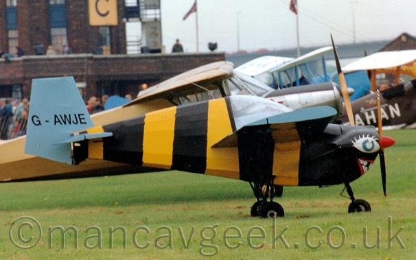 Side view of a very small, single engined light aircraft with black and yellow bands round the fuselage, pale blue wings and tail, and a mouth and eye painted on the engine cowling, vaguely resembling a bumble bee.
The registration "G-AWJE" is on the tail in black.
The plane is parked on grass, facing to the right, with several other light aircraft parked behind it.
A dark brown brick control tower fills the left of the frame, with grey skies filling the rest.