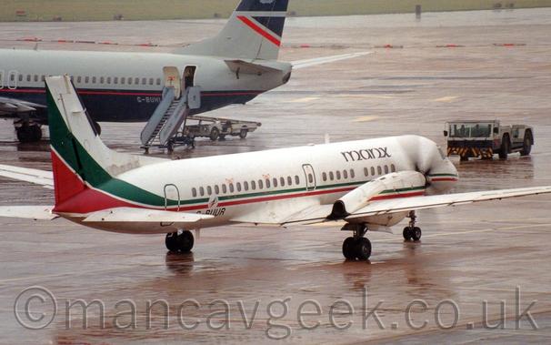 Side view of a white, twin propellor-engined airliner taxiing from left to right and slightly away from the camera, it's spinning so fast they are smeared into a blur.
There is a green and red stripe running along the body just below the passenger cabin windows, getting wider as it sweeps up into the lower part of the tail.
There are black "Manx" titles on the upper forward fuselage, and the registration "G-BUUR" on the lower rear, also in black.
Damp, grey concrete apron fills the foreground, with more in the background, as well as grey and blue rear fuselage of a jet airliner at the top left of the frame.