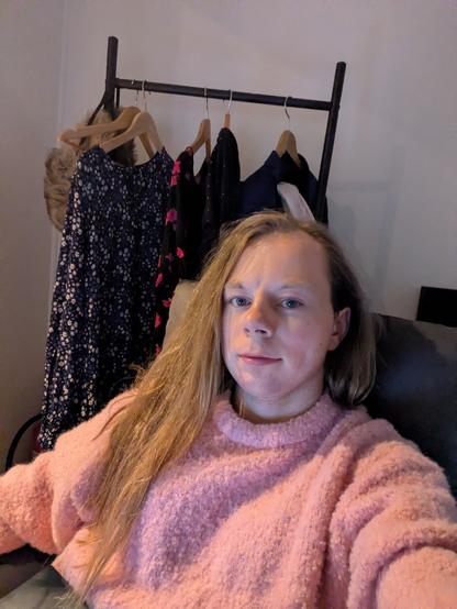 A woman sits in her apartment.

The background is simple, with a light-colored wall and ceiling. 

Some dresses hanging on a rack is visible in the background.
