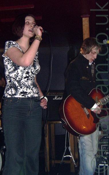 2 members of a band performing on stage, a woman with short brown hair and wearing a white top with a black flower motif and dark jeans, holding a microphone that she is singing into, and a man with short, light brown hair and wearing a dark jacket and pale blue jeans, playing a brown acoustic guitar.
Dark walls and ceiling fill the background, with a black  amplifier on a stool in the middle of the frame.