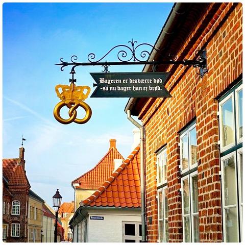 A vintage-style sign hanging from a building featuring a golden pretzel and a crown. The sign includes Danish text that translates to "The baker is dead - so now he does not bake bread." The surrounding architecture shows traditional brick buildings with sloped tile roofs.