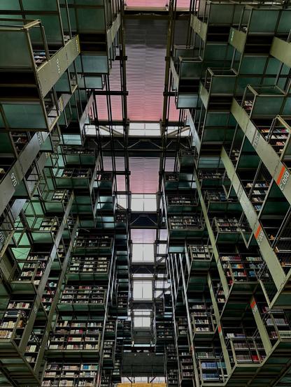 Looking up from the main floor at vast, translucent green shelving spanning multiple floors. Purple and pink light filters in across the ceiling.
