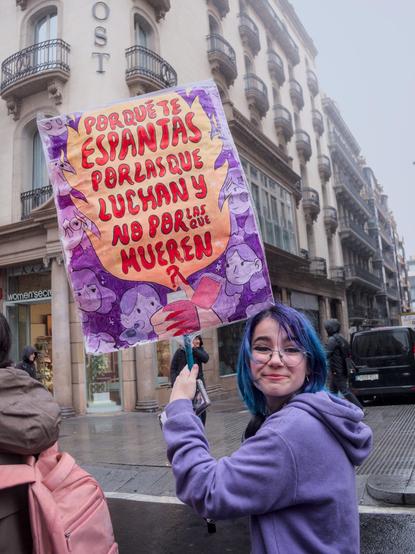 A girl with blue hair in a purple hoodie holds a sign with both hands, featuring a yellow background with red Spanish writing: "POR QUÉ TE ESPANTAS POR LAS QUE LUCHAN Y LO POR LAS QUE MUEREN" (Why are you scared of those who fight and not of those who die?). The sign's border is adorned with purple illustrations of people. 
 
Una noia amb cabell blau en una dessuadora de color porpra subjecte un cartell amb les dues mans, que presenta un fons groc amb escriptura vermella en castellà: "POR QUÉ TE ESPANTAS POR LAS QUE LUCHAN Y LO POR LAS QUE MUEREN" (Per què tens por d'aquelles que lluiten i no de les que moren?). La vora del cartell està adornada amb il·lustracions de color porpra de persones.