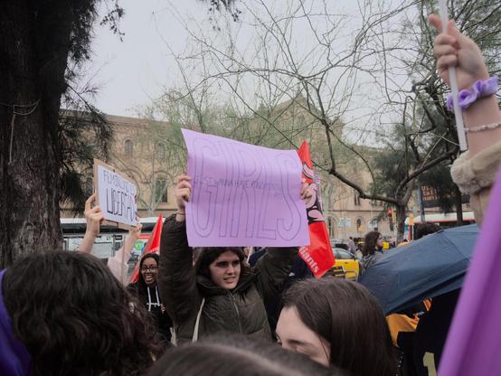 The crowd is holding signs and banners.  One girl is holding a prominent purple sign that reads "GIRLS" in large letters, accompanied by smaller text that says "JUST WANNA HAVE FUNDAMENTAL RIGHTS". On the left, another sign in Spanish "FEMINISMO ES LIBERTAD" (Feminism is liberty) can be seen.

La multitud porta cartells i pancartes. Una noia sosté un cartell de color porpra prominent que diu "GIRLS"(noies) en lletres grosses, acompanyat de text més petit que diu "JUST WANNA HAVE FUNDAMENTAL RIGHTS"(només volem drets fonamentals). A l'esquerra, es pot veure un altre cartell en castellà "FEMINISMO ES LIBERTAD" (feminisme és llibertat).