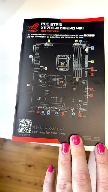The cover of the ASUS ROG STRIX X870-E GAMING WIFI motherboard Quick Start Guide. Black, glossy, but not a wasted cover page! It has a QR code in the bottom left corner for online support, and in the center, taking up most of the space, is the motherboard socket layout diagram, with colour-coded annotations and crisp white high-contrast print. The usual brand colours of black, white, and red are present and the layout is tasteful. My hot-pink polished nails are visible also, the booklet on my white desk above the hardwood floor.