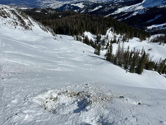 A hole blasted into the snow for avalanche control purposes showing a stable snowpack. View reveals a scenic snow-covered landscape featuring ski tracks on a snowy slope, surrounded by evergreen trees and mountain peaks in the background. A small structure is visible in the distance.