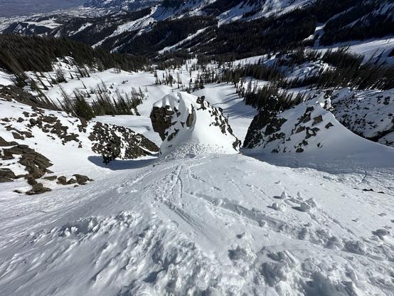 A snowy mountain landscape of steep ski slopes, rocky outcrops, and evergreen trees scattered in the distance. The scene captures a perspective looking down from a high elevation.