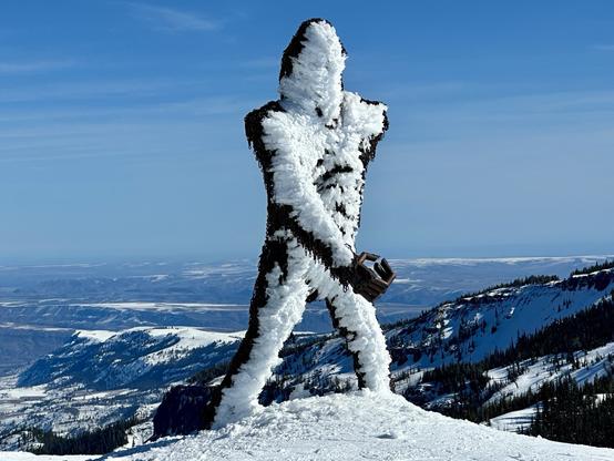 A snow-covered sculpture resembling Sasquatch stands on a snowy landscape, holding an empty clenched fist. The background features distant mountains and a clear blue sky.