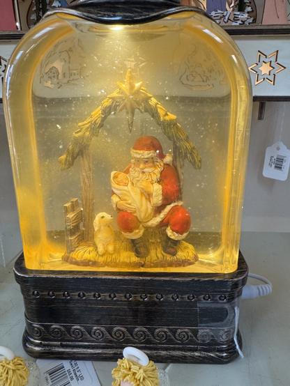 An illuminated mantelpiece glass decoration of Santa Claus holding the baby Jesus in his manger as a baby sheep looks up at them. The scene is inside a water filled glass tank.