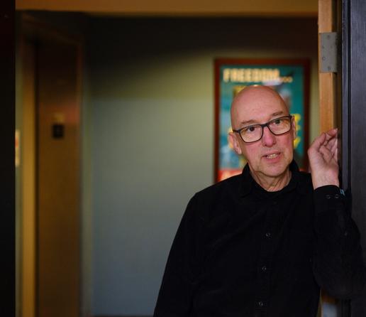 A man stands looking off to the distance, framed by a doorway.  He and a poster behind him (with the out-of-focus word FREEDOM just readable) are illuminated by late-afternoon window light.