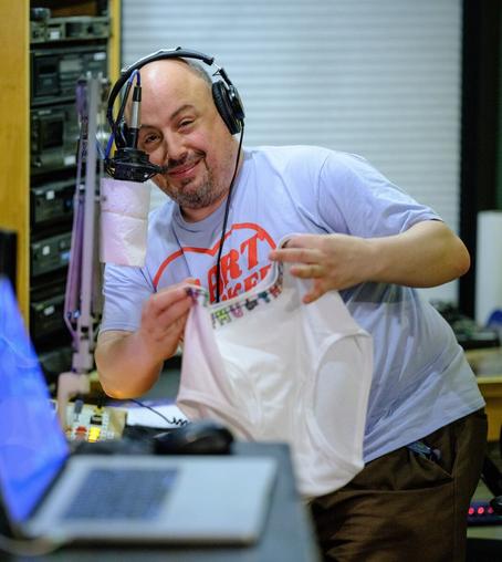 A man wearing headphones stands leaning to his right (the viewer's left) preparing to unfurl some white underpants