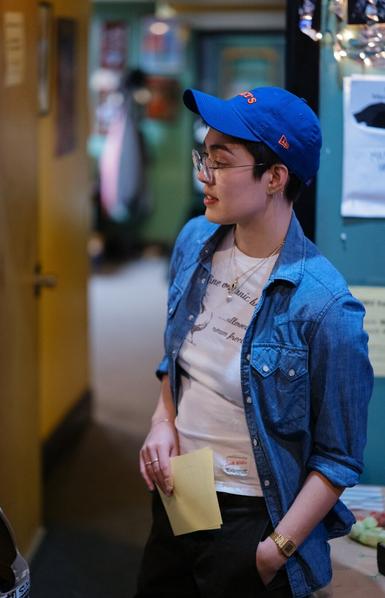 A woman in a baseball cap and jean jacket stands, one hand in trouser pocket; that wrist sports a small gold-tone digital watch.
