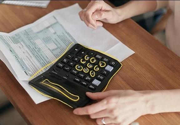 Es veu una calculadora sobre una taula i una persona que està fent comptes amb ella i té uns papers al costat.