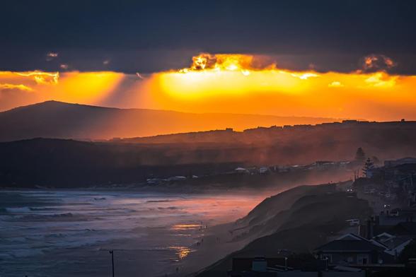 The image shows a dramatic coastal scene at sunset or sunrise, with a beach in the foreground and ocean waves. The sky is filled with dark clouds, but there's a striking orange and yellow glow from the sun, casting warm light across the landscape. Hills or mountains can be seen in the background, and there are some buildings along the shore. The contrast between the dark clouds and the bright sunlight creates a vivid and captivating atmosphere.