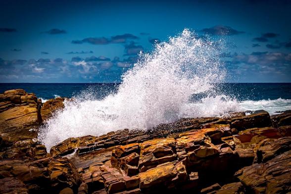 The image shows waves crashing against rocky formations on a coast. The water is splashing upwards, creating a dramatic display against the vibrant blue of the ocean and sky. The rocks in the foreground appear rugged and weathered.