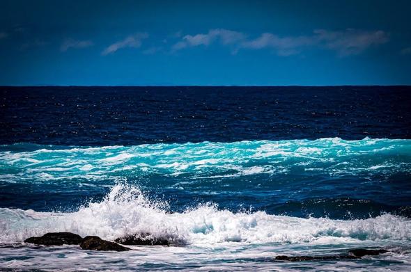 The image shows a view of the ocean, featuring waves crashing against rocks. There is a clear distinction between the foamy, breaking waves in the foreground and the deeper blue water in the background. The sky is visible above the horizon.