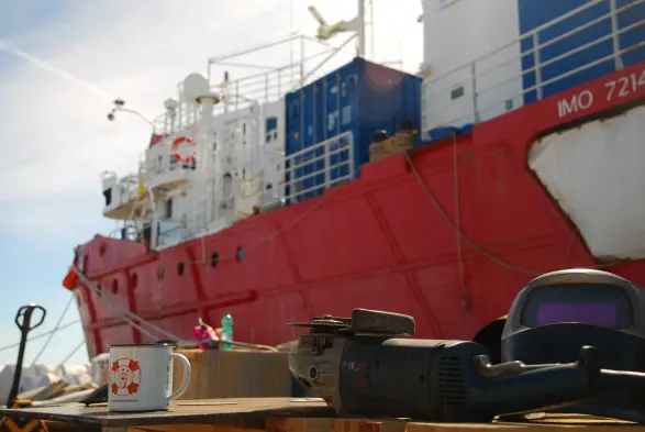 bild im querformat: im vordergrund ein großer winkelschleifer, ein schweißhelm und eine tasse der ngo aurora suport.
im huntergrund das rettungsschiff sea eye 4 am pier