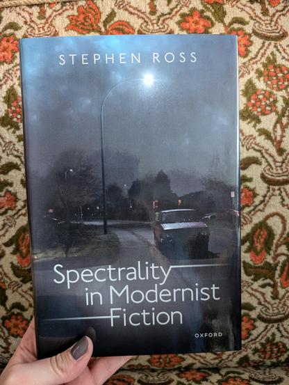 Holding the book "Spectrality in Modernist Fiction" by Stephen Ross in front of a chair with an orange flower pattern