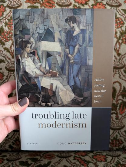Holding the book "Troubling Late Modernism" by Doug Battersby in front of a chair with an orange flower pattern
