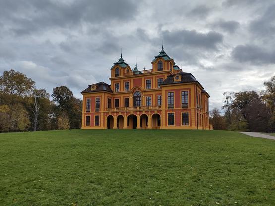 Das Bild zeigt ein eindrucksvolles, barockes Schloss mit einer leuchtend gelb-orangefarbenen Fassade und grünen Dächern. Das Gebäude ist symmetrisch gestaltet und besitzt mehrere Türme mit kleinen Fenstern. Die Architektur ist durch große Fenster, dekorative Balustraden und eine imposante Arkadenreihe im Erdgeschoss geprägt. Das Schloss steht auf einer sanft ansteigenden, grünen Wiese, die sich vor dem Gebäude erstreckt. Im Hintergrund sind herbstlich gefärbte Bäume zu sehen, die das Schloss umgeben. Der Himmel ist bewölkt, was eine dramatische Lichtstimmung erzeugt, während die Farben des Schlosses im Kontrast zu den dunklen Wolken besonders kräftig erscheinen. Ein Weg führt seitlich am Schloss vorbei