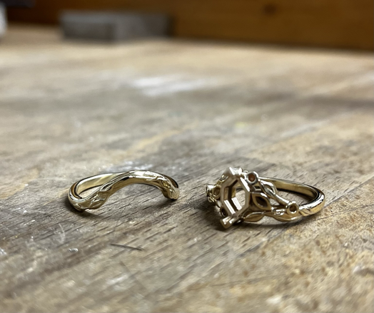 A set of rings in yellow gold on a wooden light coloured table. The engagement ring has a leaf pattern and no stone set yet into the main setting, a curved wedding band wifh leaf pattern is placed next to it the curved side upwards.