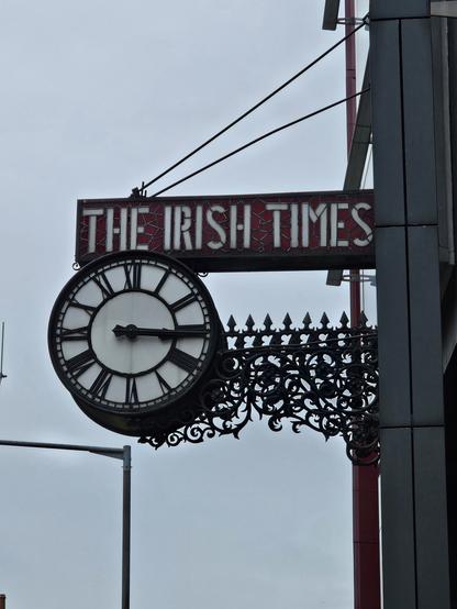 Photograph of a clockface on the side of the Irish Times building. 'The Irish Times' is writtenr in white stained glass on a red background above thd clock. The clock has riman numeraks, where 4 is 4 parallel lines, not IV