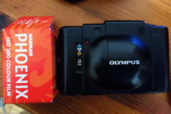 Farbfoto, das eine kompakte Filmkamera von ca. 1979 zeigt - die Olympus XA. Links daneben eine rote Schachtel mit einem Phoenix-Farbfilm von Harman.