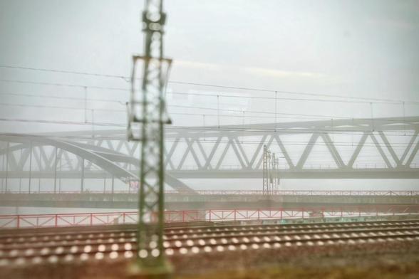Eine neblige Szene mit einer Eisenbahnbrücke im Hintergrund, mit Stromleitungen und Bahngleisen im Vordergrund. Die Sichtbarkeit ist aufgrund des Nebels gering, was dem Bild eine gedämpfte und atmosphärische Qualität verleiht.

A foggy scene featuring a railway bridge in the background, with power lines and train tracks in the foreground. The visibility is low due to mist, giving a muted and atmospheric quality to the image.
