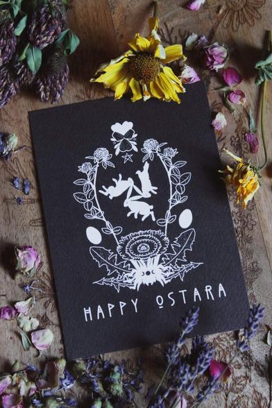 Das Bild zeigt eine schwarze Karte mit weißen Illustrationen und dem Text „HAPPY OSTARA“ unten. Um die Karte herum liegen verschiedene getrocknete Blumen und Kräuter, darunter Klee, Lavendel und Rosen. Auf der Karte sind drei Hasen, Eier und florale Elemente in einem symmetrischen Design abgebildet. Die Karte liegt auf einer Holzoberfläche mit weiteren floralen Gravuren.