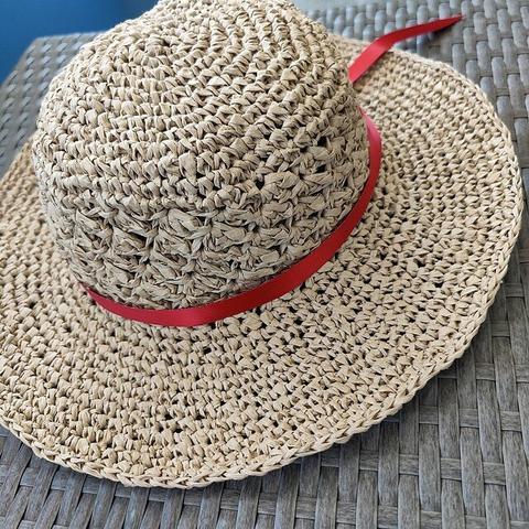 A crocheted hat made with raffia yarn is shown. The hat is beige in color and features a wide brim. It is adorned with a red ribbon tied around the base of the crown. The hat is placed on a woven dark-grey surface.
