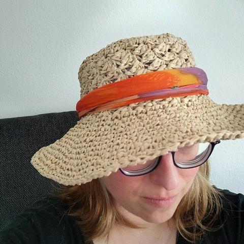 A person wearing a crochet straw hat with a wide brim and a colorful orange, yellow and purple scarf wrapped around the base of the hat. The background shows a white wall and a dark-colored couch. The face of the person is partly obsucred by the hat so that only the lower half of the face is visible.