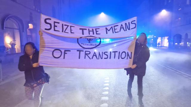 In a demonstration at night, two TransHub members hold a purple and black banner that reads "Seize the means of transition".