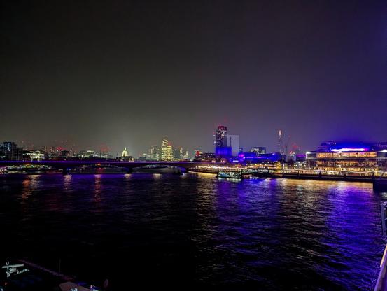 night scene of a wide river with a cityscape and a road bridge. the buildings on the right bank and the parapet of the bridge are lit in purple which is reflected in the water surface