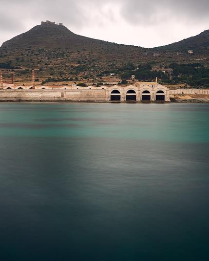 In the foreground you see the seawater inside the harbor like a plane due to the long exposure. The seawater is turquoise with dark patches. At the end of the harbour the ruins of an old tuna factory are seen. Behind the ruins small mountains or hills are seen with the ruins of a castle on top of the largest one. Above a dark blue sky with only patches of white clouds are seen.