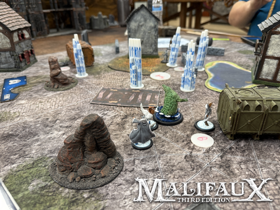 A tabletop miniatures game in progress. In the foreground, amidst terrain, are (seen from the back) minis for Rasputina, a human winter witch; a white fur-cloaked Silent One, a member of the cult of December; Bashe, a giant alligator; and Sightless Snow, an ally of Rasputina's. Past them are a number of blue-runed ice pillars, and among them are Leviticus' haplessly ensnared crew.