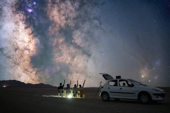 Pexels photo shows a desert scene at night, under a lit-up blanket of stars & the Milky Way. At center bottom are 4 figures seated on plastic chairs, facing away from us and toward the celestial display. To the right is a sporty white hatchback car with its rear hatch open and a camera tripod just outside. Possibly young men, the 4 figures each have their right arm in the air, joyfully saluting the cosmos. Mood is one of celebrating friendship and the grandeur of the night sky. 
Captioned, "Stargazers Under the Milky Way in Iran Desert". 

Semnan, Semnan Province, Iran. 
Dated 2024 Sep 11, by atiabii.
www.Pexels.com/photo/stargazers-under-the-milky-way-in-iran-desert-28351764/ 

www.KronoMoon.org 🏳️‍🌈☮️ 
#KronoWatch
