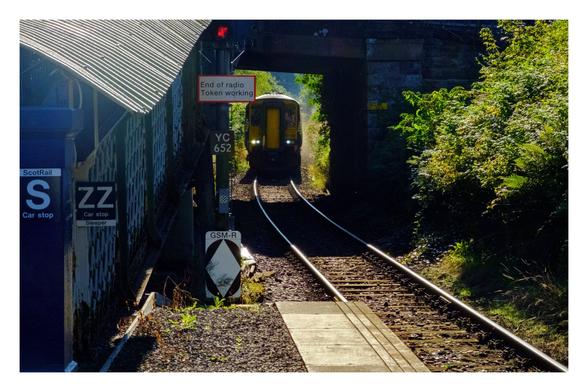 Foto im Querformat. Blick entlang einer Eisenbahnschiene vom Gleis aus. Ein gelber Zug fährt gerade ein. Bevor er am Gleis ankommt, muss er noch durch eine kurze Unterführung fahren. Links ist der überdachte Zugang zum Gleis. Daran sind verschiedene Schilder. Daraus ist erkennbar, dass wir uns in Schottland befinden. Es ist ein sonniger Tag.