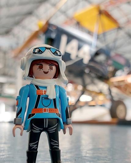 Mulher playmobil com capacete antigo branco de aviador e oculos, vestida de azul. Por trás um avião biplano